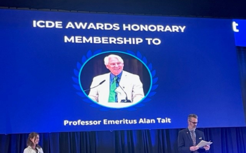 The backdrop at an awards ceremony shows a slide. Copy reads: ICDE Awards Honorary Membership to Alan Tait, with a photo of Alan in the centre