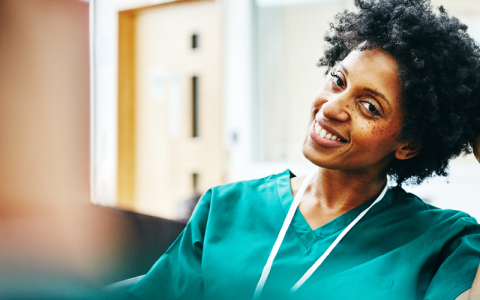 A relaxed female nurse in turquoise scrubs looks at the camera and smiles.
