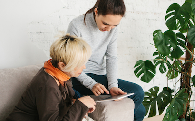 A young woman sitting on the arm of a sofa shows an older woman something in a booklet. Both are dressed casually.