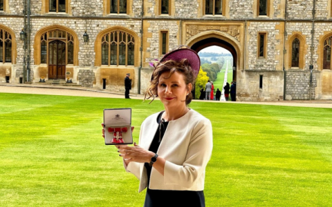Jane stands outside Windsor Castle. She's dressed very smartly and is displaying her MBE in its presentation case.