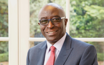 portrait of Professor Kwame Akyeampong. He is sitting in front of a window, facing the camera and smiling. He is wearing a red tie and glasses. 