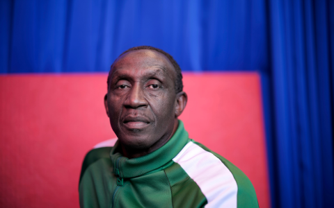 Linford Christie is wearing a green tracksuit top with a white stripe down the sleeve.  He is staring into the camera with a serious expression on his face 
