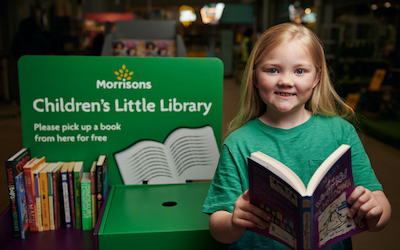 A young (junior aged) girl in a green top looks at the camera, holding open a book. She is standing beside a stand advertising the Morrison's Children's Little Library.