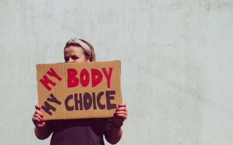 Woman holding a homemade cardboard sign saying my body, my choice. It's partially covering her face. 