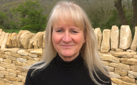 Portrait photograph of Professor Victoria Cooper. She has long blonde hair and is wearing a black top. She is stood in front of a sandstone wall in a field. 