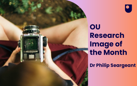 A graphic showing a photo that looks down on a girl sitting on grass holding a camera. The words to the right of the graphic read: OU Research Image of the Month Dr Philip Seargeant