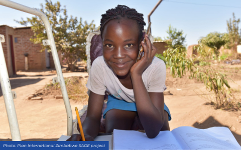 Zimbabwean schoolgirl on mobile phone whilst writing in notebook outdoors.