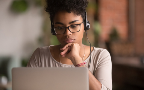 A young black woman sits looking thoughtfully at a laptop screen. She wears glasses and headphones and is resting her chin on her hand.