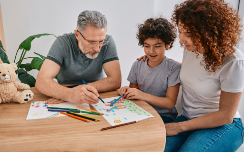A child, male adult and female adult sitting around a table. On the table is a children's drawing which the child and the adult male have pens poised over.  A teddy bear sits on the table to the left of the three individuals.