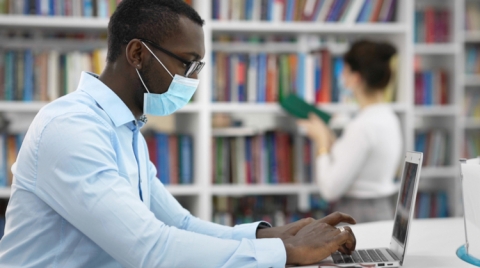 A male student wearing a face mask sits at a laptop in a library. A woman (out of focus) looks through books in the background.