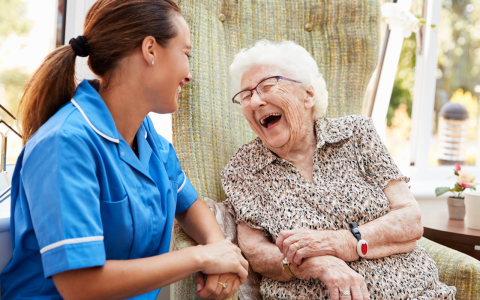 A carer wearing a blue uniform is sitting talking to an elderly lady. The elderly lady is laughing joyously