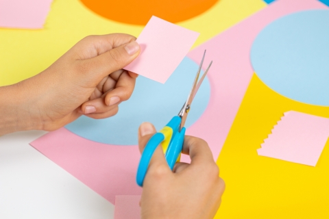 This image shows a person's hands holding a pair of scissors and pink paper with colourful paper in the background.