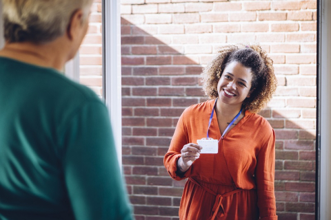 A photograph of a Social Worker smiling and showing their ID badge to a person at a front door.
