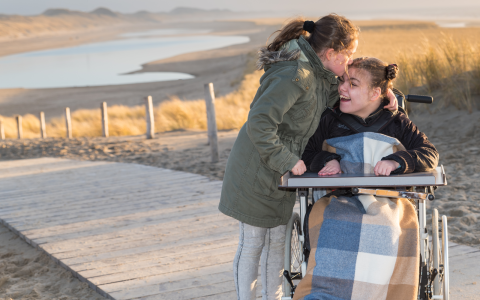 A young disabled girl in a wheelchair at the beach with her sister. The girl in the chair is smiling while her sister kisses her head