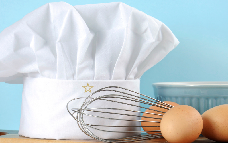 A star baker chef's hat with a whisk, mixing bowl and eggs.