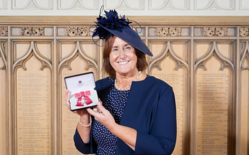 Dr Janice Gorlach is pictured holding her MBE. She is dressed smartly in navy blue and is wearing a hat. 