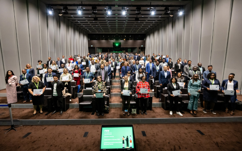 The FIFA graduates stand next to their seats in a lecture room, all holding their diplomas.