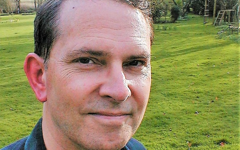 close up portrait of Jonathan Yonge. He's wearing a blue shirt and is in a garden with a green lawn in the background. 