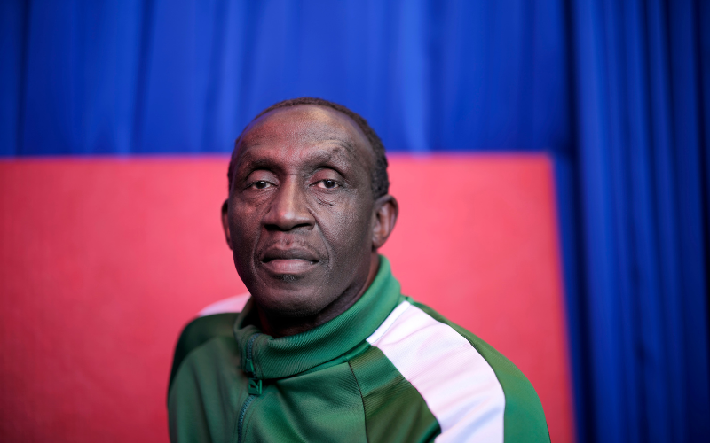 Linford Christie is wearing a green tracksuit top with a white stripe down the sleeve.  He is staring into the camera with a serious expression on his face 