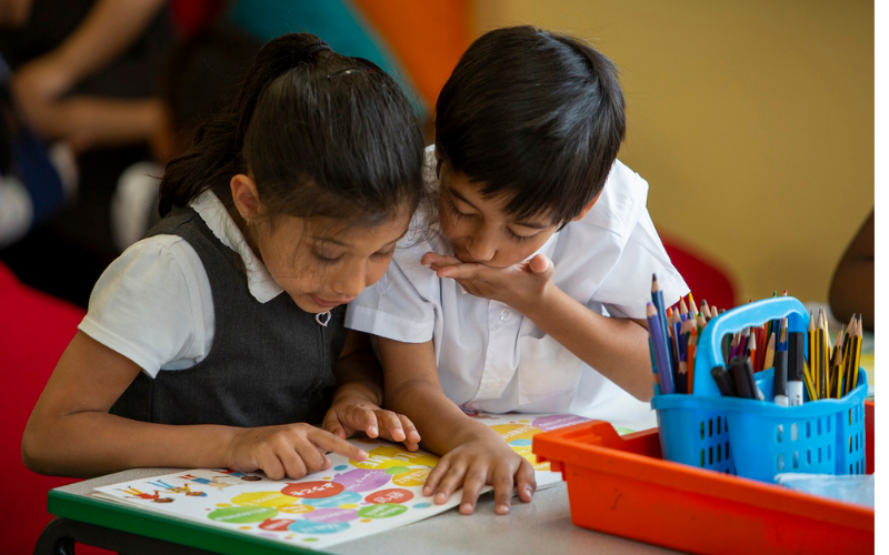A primary school aged boy and girl intently examine a reading book together, in a classroom setting.