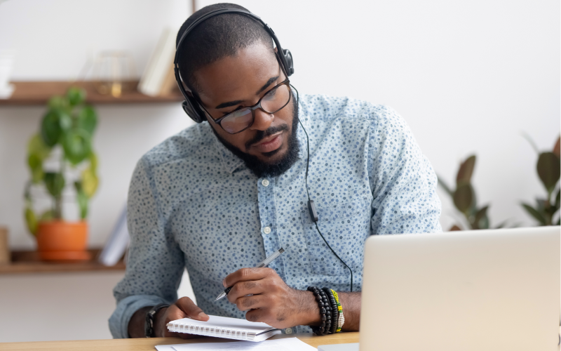 A man is sitting at a laptop computer, staring intently at the screen. He's wearing a headset, and is taking notes on a reporters notepad. 